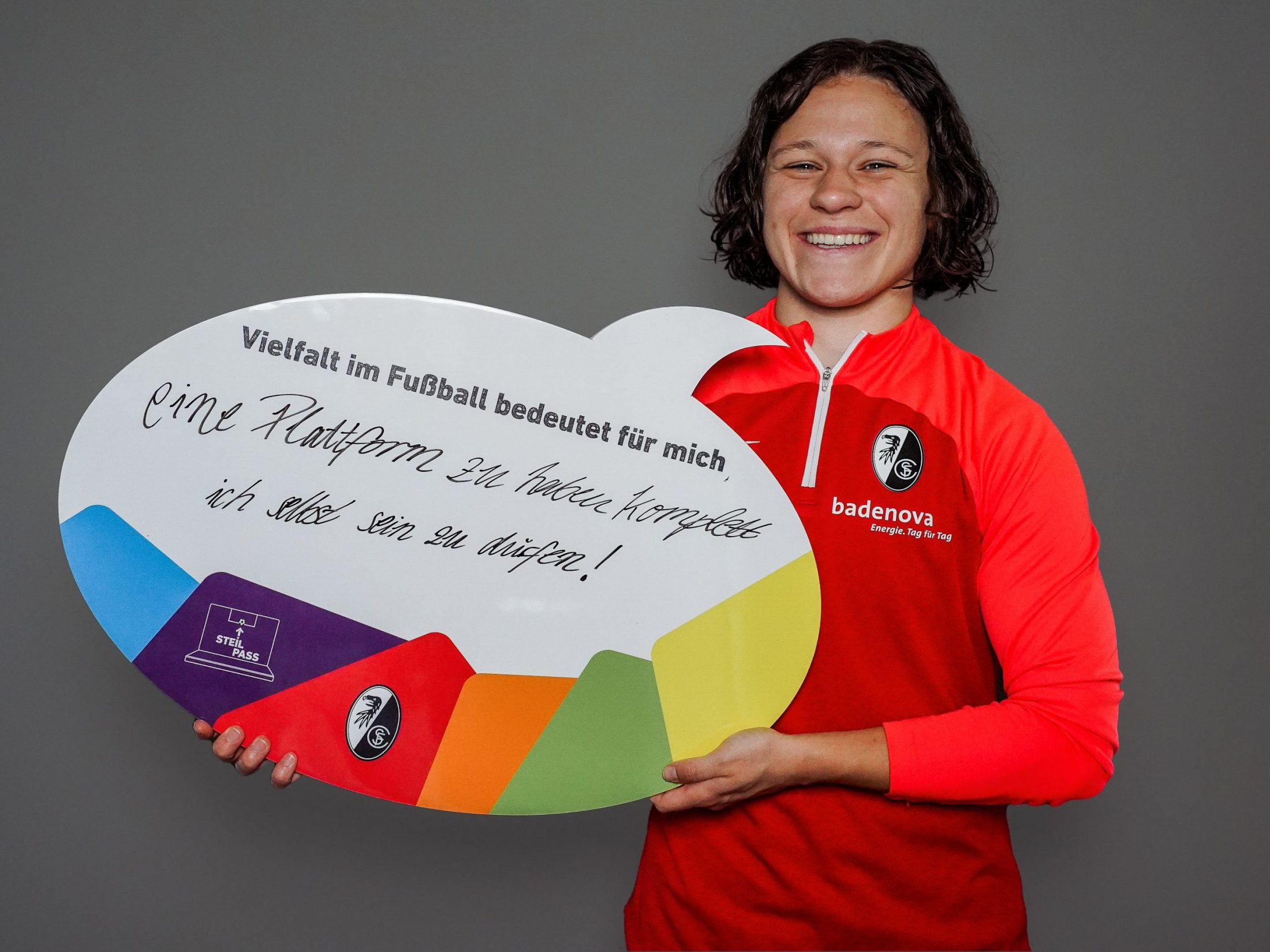 Weiße, weiblich gelesene Person mit einer SC Freiburg-Jacke hält eine Sprechblasenschild hoch. "Vielfalt im Fußball bedeutet für mich, eine Plattform zu haben - komplett ich selbst sein zu dürfen!", steht darauf geschrieben.