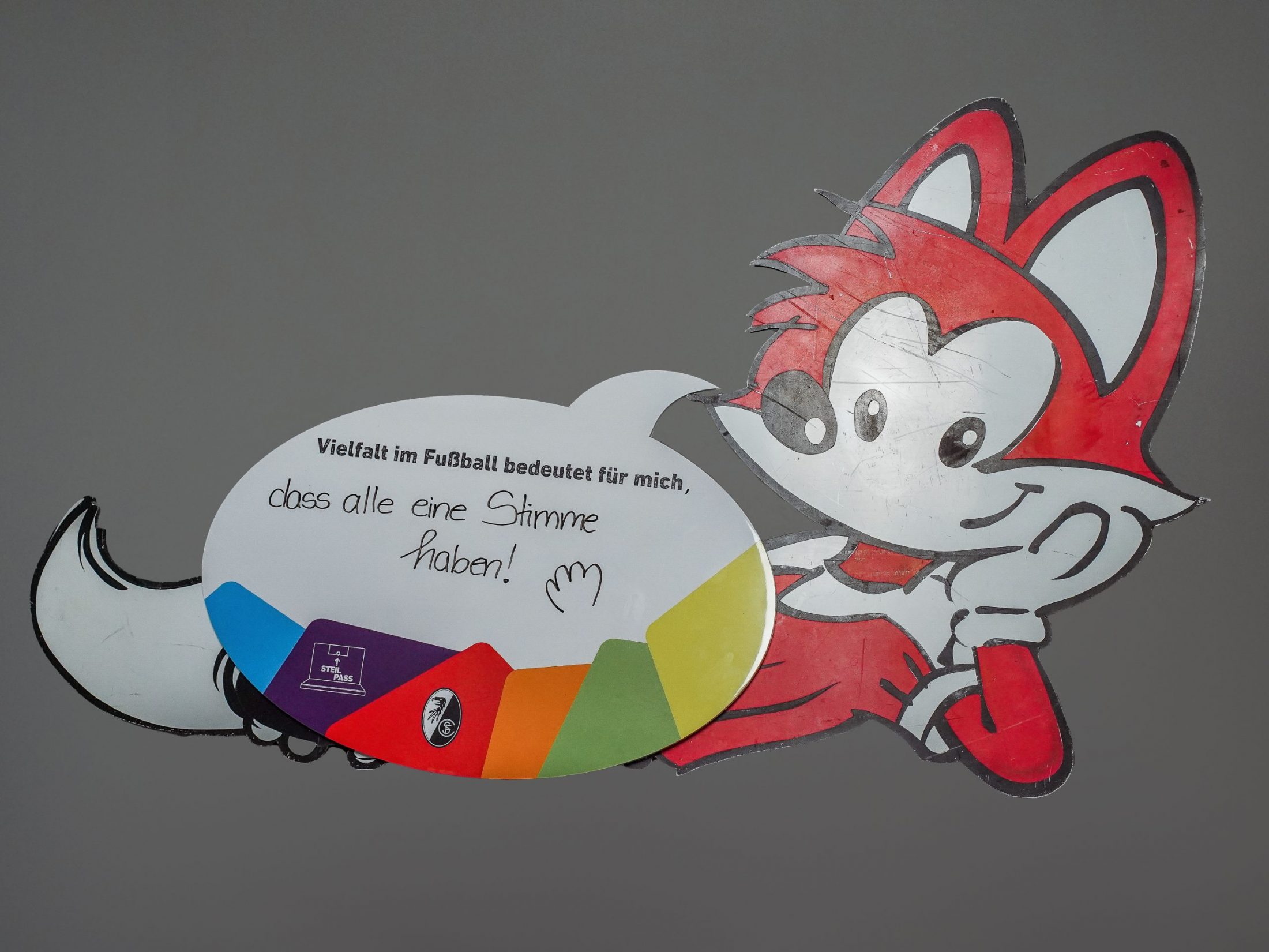 Eine rote Fuchs-Figur hält eine Sprechblasenschild . "Vielfalt im Fußball bedeutet für mich, dass alle eine Stimme haben!", steht darauf geschrieben.