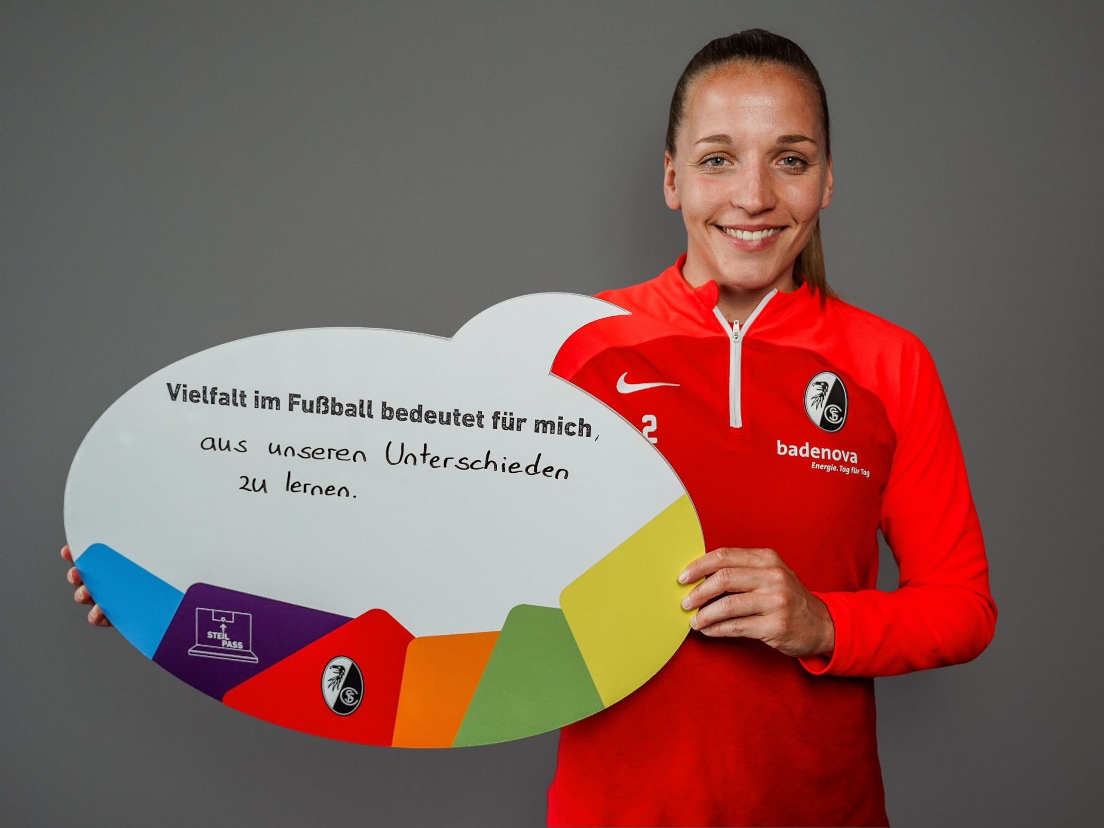 Weiße, weiblich gelesene Person mit einer SC Freiburg-Jacke hält eine Sprechblasenschild hoch. "Vielfalt im Fußball bedeutet für mich, aus unseren Unterschieden zu lernen", steht darauf geschrieben.